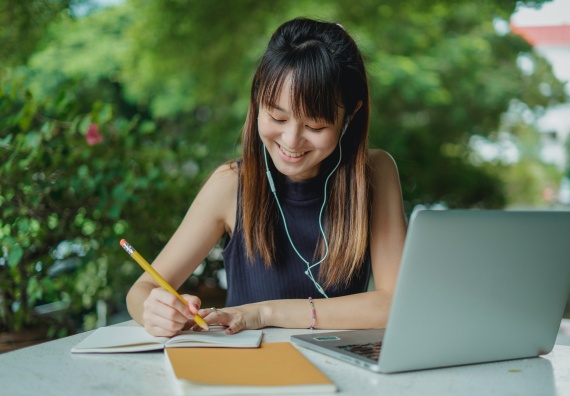 Alt=" A chinese lady seated and smiling while taking notes and writing down from a laptop as to a booklet"